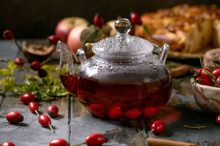 Winter hot beverage Rosehip berries herbal tea in glass teapot standing on old wooden plank table with wild autumn berries and apple cake around. Healthy hot drink.の写真素材