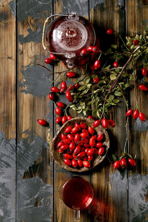 Rose hip berries herbal tea in glass teapot and cup standing on old wooden plank background with wild autumn berries around. Winter hot beverage. Flat layの写真素材