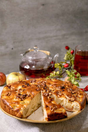 Homemade apple and walnuts cake Charlotte on plate with fresh gardening apples, nuts, rose hip tea around on grey linen table cloth. Autumnal cozy baking.の写真素材