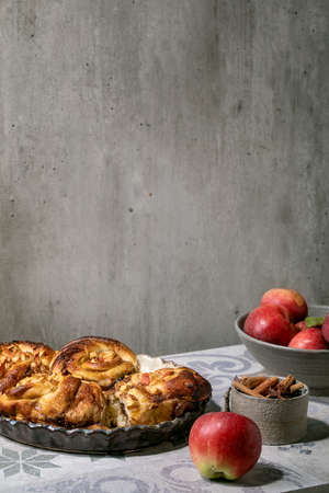 Sweet apple sticky cinnamon rolls buns in round baking tray standing with gardening apples and cinnamon sticks on ornate ceramic tile table. Traditional home bakery. Copy spaceの写真素材