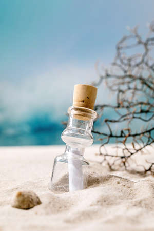 Glass bottle with message paper inside on summer sand sea beach with waves and coral branch at background. Help SOS fate message concept.の写真素材