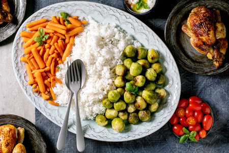 Dinner table with grilled chicken, rice and vegetables baked brussel sprouts, baby carrot on big oval dish in center and guest plates on blue linen tablecloth over grey background. Flat lay.の写真素材