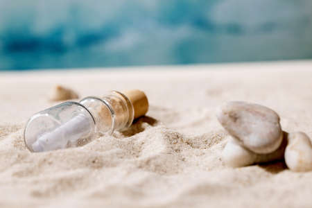 Glass bottle with message paper inside lying on summer sand sea beach with waves at background. Copy space creative layout. Help SOS fate message concept.の写真素材