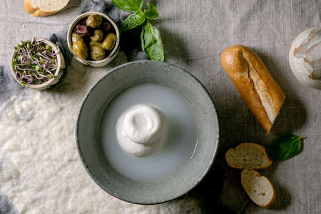 Traditional italian burrata knotted cheese in grey ceramic bowl on table. Bread, olives, green salad...