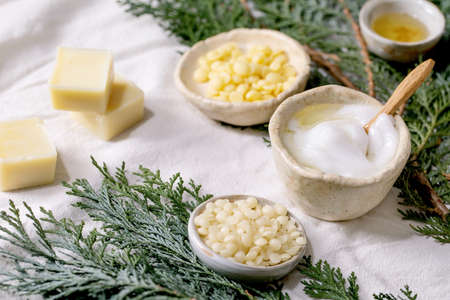 Ingredients for makes home made natural cosmetic. Face cream in progress, essential oils, cocoa butter, wax and soap in ceramic bowls on white linen cloth with thuja branches. Eco friendly DIY.の写真素材