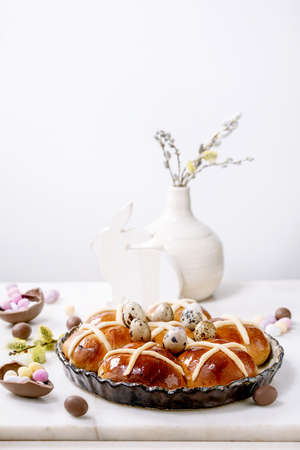 Homemade Easter traditional hot cross buns in ceramic dish with blossom flowers, chocolate candy and quail eggs on white marble table. Easter holiday baking and decorの写真素材