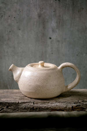 Craft white speckled handmade ceramic teapot kettle for tea ceremony standing on old wooden shelf with grey wall at backgroundの写真素材