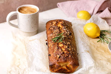 Homemade lemon pound cake with nuts and rosemary on crumpled baking paper, serving with fresh lemon and coffee mug on white marble table.の写真素材