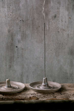 Fuming incense stick in hand crafted gray ceramic incense holder on wooden table with grey wall at...