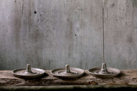 Set of hand crafted gray ceramic incense holder with fuming incense stick on wooden table with grey wall at background.の写真素材