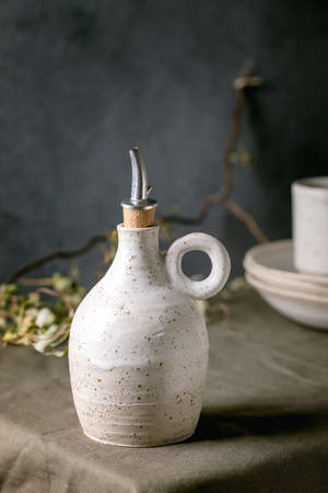 White speckled craft ceramic olive oil or vinegar bottle dispenser standing on dark green linen tablecloth with stack of plates. Rustic style, coly spaceの写真素材
