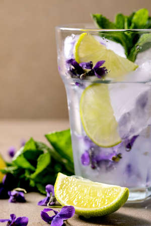 Transparent glass of floral lemonade soda cocktail with mint, violets flower ice and lime slices standing on brown table. Cold summer refreshment drinkの写真素材