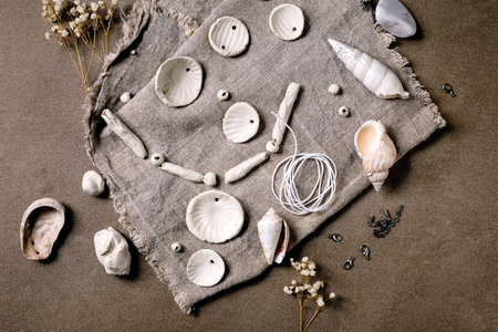 Ceramic workshop idea: handcrafted beads from clay or porcelain. Flat beads of raw white porcelain on rough fabric. Small white flowers, jewelry findings, sea shells. Dark background, top view.の写真素材