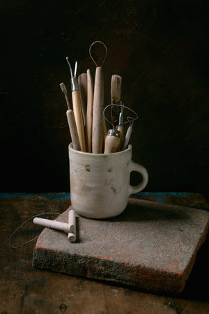 Set of clay-working tools. Old pottery tools for trimming and forming clay items in ceramic mug, wire tool, cutting string on aged wooden table with dark background.の写真素材