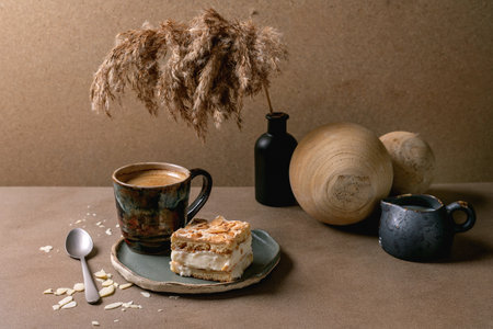 Homemade Norwegian sweet cake Verdens Beste Kake with custard, meringue, and almonds, one square piece on plate on beige table with coffee cup, spoon, milk jug, reed in bottle, beige wall.の写真素材