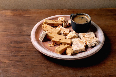 Traditional Spanish dessert classic soft orange turron and crunchy almond turron, cut into pieces on rustic clay plate. Espresso in black cup, wooden table, copy space.の写真素材