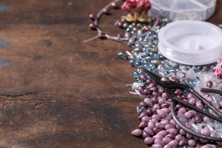Handmade jewelry making. Creating beaded necklace with glass beads and components in ceramic bowl. Scissors, jewelry findings, spring flowers on dark wooden background. DIY hobby concept. Copy spaceの写真素材