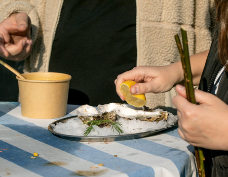 Street food festival Europe. Woman squeezing lemon over fresh oysters on ice plate. Traditional seafood delicacy served outdoor table. Gourmet street cuisine experienceの写真素材