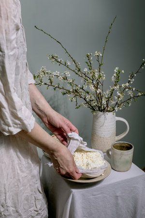 Woman in white linen clothing serves fresh homemade cottage cheese wrapped in cloth. Ceramic pitcher with blooming white spring branches and tea cup on white tablecloth. Rustic elegant setting.の写真素材