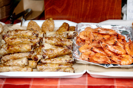Delicious homemade spring rolls with meat filling served alongside cooked prawns in foil tray. Party food ready for serving on white plates against wooden background.の写真素材