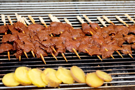 Marinated beef skewers on wooden sticks grilling on barbecue grill with sliced yellow potatoes. Traditional street food summer cookout outdoor cooking.の写真素材