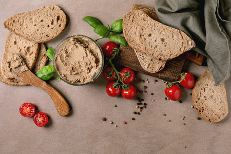 Homemade liver pate in glass bowl with whole grain bread slices fresh cherry tomatoes basil leaves on wooden cutting board. Rustic breakfast setup on beige background.の写真素材