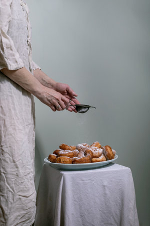 Woman wearing linen clothes sprinkling powdered sugar through sieve onto fresh homemade donuts served on plate against minimalist background.の写真素材