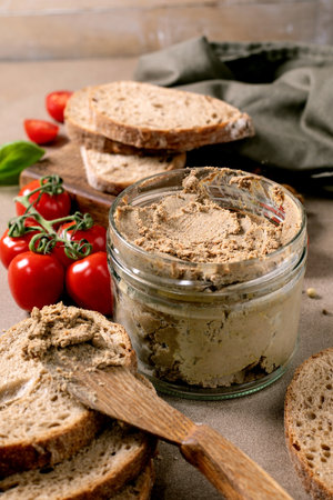 Homemade meat pate in glass jar served with fresh whole grain bread slices cherry tomatoes and basil leaves. Rustic wooden spoon and cutting board.の写真素材