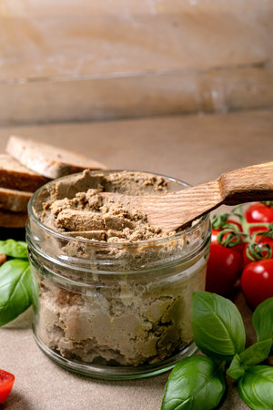 Closeup of glass jar with homemade liver pate wooden spoon fresh basil leaves cherry tomatoes and bread slices on rustic background.の写真素材