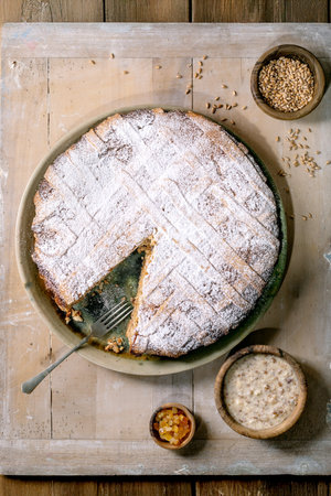 Rustic homemade cake Pastiera Napolitano dusted with powdered sugar on plate slice cut out accompanied by wooden bowls of seeds porridge dried fruits on wooden table background. Top view.の写真素材