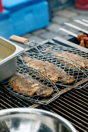 Fresh fish grilling process on barbecue grill using metal mesh basket. Summer outdoor cooking with kitchen utensils visible around grill.の写真素材