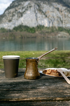 Coffee break with copper cezve pastry snack Austrian strudel on wooden table against beautiful mountain lake landscape. Perfect outdoor meal with breathtaking nature view.の写真素材