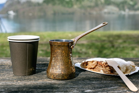 Traditional copper coffee pot with disposable cup and Austrian strudel on wooden table outdoors near lake. Morning breakfast picnic setting lakeside.の写真素材