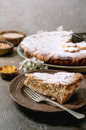 Slice of homemade pie Pastiera Napolitano dusted with powdered sugar served on wooden plate. Whole tart with ingredients in background on rustic table.の写真素材