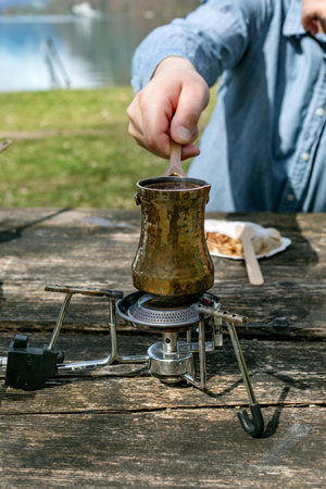 Person brewing traditional Turkish coffee in copper cezve on portable camping stove. Outdoor picnic near lake wooden table surface. Closeup hand stirring coffee.の写真素材