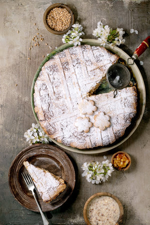 Traditional homemade pie Pastiera Napolitano with powdered sugar served with slice on plate. Decorated with white flowers grains dried fruits and strainer. Rustic setup on gray concrete background.の写真素材