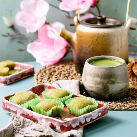 Asian traditional rice dessert sweet green matcha mochi in pink plate with cup of frothed matcha tea on blue table. Old teapot and spring pink flowers at background.の写真素材