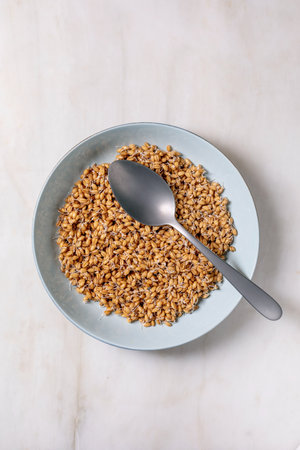 Top view of sprouted wheat grains in light blue ceramic bowl with metal spoon on white marble...