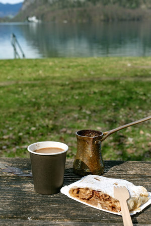 Coffee traditional cezve pot and Austrian strudel pastry on wooden table by mountain lake. Perfect outdoor picnic breakfast with scenic nature view background.の写真素材
