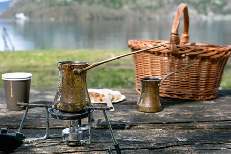 Traditional brass cezve pot brewing coffee on portable stove outdoors. Picnic setting with wicker basket and Austrian strudel wooden table lake background.の写真素材