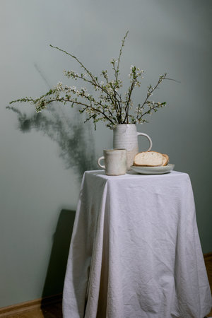 Minimalist still life with white ceramic pitcher filled with spring blossoms mug and bread slice on plate, all arranged on table covered with white linen tableclothの写真素材