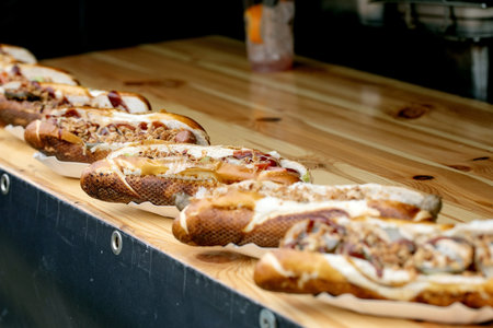 Row of delicious loaded hot dogs with various toppings in paper holders displayed on wooden counter at street food festival. Gourmet sausages with melted cheese bacon bits and condiments.の写真素材