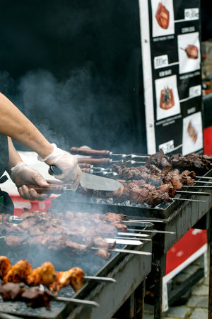 Chef wearing gloves grilling meat skewers at outdoor street food market. Smoke rising from barbecue grill with menu board display in background.の写真素材