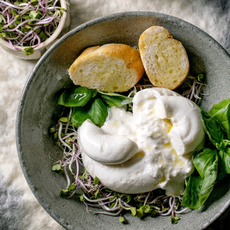 Traditional Italian burrata knotted cheese salad in gray ceramic bowl on table. Sliced bread, olives, green sprouts around. Healthy mediterranean dinner. Top viewの写真素材
