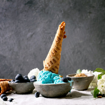 Homemade turquoise ice cream with waffle cone in ceramic bowl. Blueberries and white flowers around. Standing gray linen table cloth. Trendy homemade summer dessertの写真素材