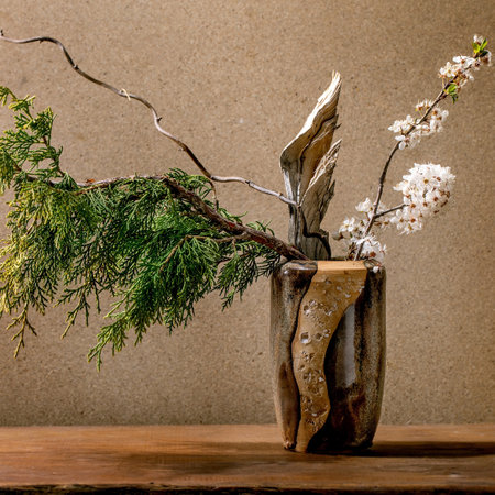 Beautiful spring ikebana. Floral arrangement with spring blooming white flowers, thuja branch and piece of wood in brown ceramic vase, standing on wooden table. Japanese style home decorの写真素材