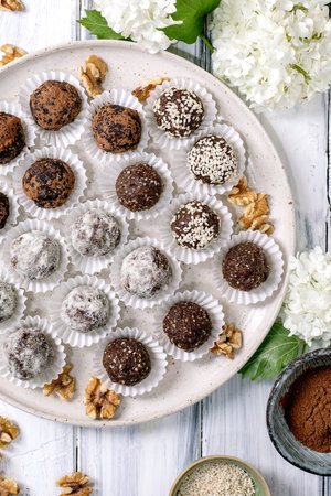 Assorted homemade chocolate raw vegan balls truffles in paper cups on ceramic plate. Different coatings: cocoa powder powdered sugar sesame seeds. Decorated with walnuts and white flowers.の写真素材