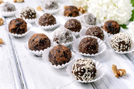 Assorted homemade chocolate raw vegan balls truffles with different toppings in paper cups on white wooden background decorated with white flowersの写真素材