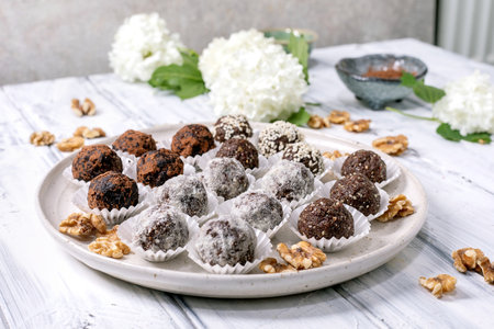 Homemade chocolate raw vegan balls truffles variety with different toppings on white ceramic plate. Dusted with cocoa powder sesame seeds. Decorated with walnuts and white flowers.の写真素材