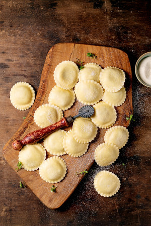 Homemade ravioli pasta arranged on rustic wooden cutting board with vintage pasta cutter dusted with flour on dark wood backgroundの写真素材